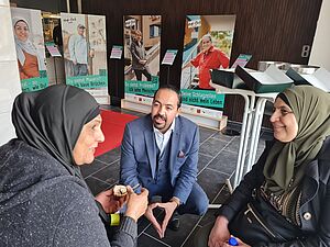 Zwei weibliche Personen mit Kopftuch, dunkel gekleidet und eine männliche Person im Anzug sitzen im freundlichen Gespräch miteinander. Hinter ihnen eine Roll-up-Ausstellung mit Portraits von Personen. Die Roll-ups sind mit kleinen Tafeln jeweils danebenstehend beschriftet.