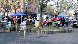 Menschen sitzen und stehen an Tischen unter blauen Pavillons, während eine Freiluft-Ausstellung mit Fotos und Bildern im Vordergrund zu sehen ist.