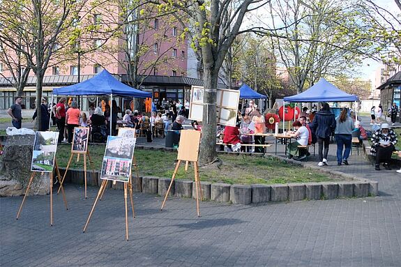 Menschen sitzen und stehen an Tischen unter blauen Pavillons, während eine Freiluft-Ausstellung mit Fotos und Bildern im Vordergrund zu sehen ist.