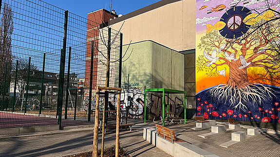 Sitzbänke und neue Bäume stehen vor dem Bolzplatz mit rotem Belag. 