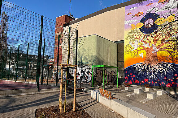 Sitzbänke und neue Bäume stehen vor dem Bolzplatz mit rotem Belag. 