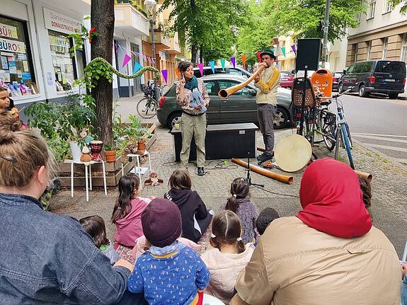 Kinder und Erwachsene sitzen auf dem Gehweg vor dem QM-Büro Donaustraße Nord und verfolgen eine Didgeridoo-Aufführung mit Musik und Puppen.