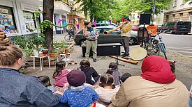 Kinder und Erwachsene sitzen auf dem Gehweg vor dem QM-Büro Donaustraße Nord und verfolgen eine Didgeridoo-Aufführung mit Musik und Puppen.