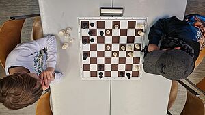 Aufnahme von oben auf ein Schachbrett, das auf einem Tisch liegt. Auf beiden Seiten sitzen die Spielenden, es sind zwei Kinder.