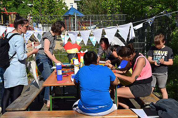 Mehrere Teilnehmende sitzen um einen Tisch und gestalten gemeinsam Wimpelketten. 