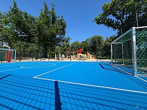 Ein blaues Spielfeld mit zwei Fußballtoren und Spielfeldmarkierungen. Im Hintergrund ein Spielplatz und Bäume. Der Himmel ist blau.