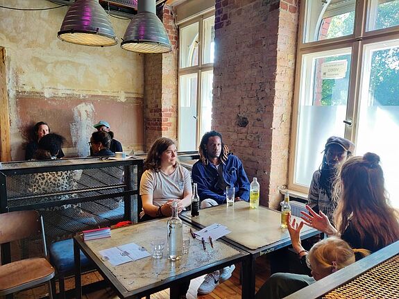 An zwei Tischen sitzen Gruppen und reden über die Gestaltung des Panafrican Festivals.