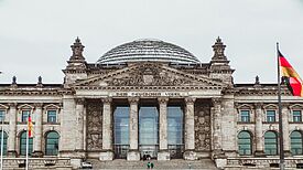 Das Reichstagsgebäude in Berlin mit der Aufschrift „Dem Deutschen Volke“ und der gläsernen Kuppel, flankiert von zwei deutschen Flaggen.