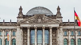 Das Reichstagsgebäude in Berlin mit der Aufschrift „Dem Deutschen Volke“ und der gläsernen Kuppel, flankiert von zwei deutschen Flaggen.