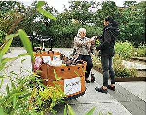 Eine ältere Frau mit Rollator spricht lächelnd auf einem Weg mit einer jüngeren Frau, die ihr etwas gibt. Neben ihnen ein Lastenrad mit der Aufschrift „Redemobil“. In Vorder- und Hintergrund Sträucher, Bäume und bepflanzte Beete.