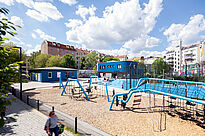 Im Vordergrund steht ein großes Kletter- und Spielgerät auf einer mit Holzschnitzeln bedeckten Fläche. Dahinter liegen ein blauer Vorplatz, ein eingezäunter Bolzplatz und das Blueberry-Gebäude, das von mehrgeschossigen Wohnhäusern umgeben ist.