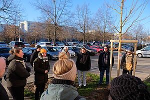 Eine Gruppe von zehn Erwachsenen steht um einen neu gepflanzten Baum auf einem kleinen Stück Rasen. Angrenzend ein Parkplatz mit dicht parkenden Autos, im Hintergrund Wohnungsneubauten