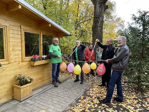 Mehrere Personen schneiden dekorierte Luftballons an einem Zweig vor einer neuen Holz-Blockhütte im Volkspark Lichtenrade durch.
