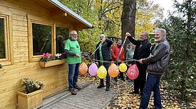 Mehrere Personen schneiden dekorierte Luftballons an einem Zweig vor einer neuen Holz-Blockhütte im Volkspark Lichtenrade durch.