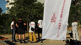 Mehrere junge Menschen, Rückenansicht, stehen draußen vor einem Baum und sprechen miteinander. Sie tragen T-Shirts mit der Aufschrift „team“. Angeschnitten im Bild sind Sportgeräte. Im Vordergrund ein wehendes Banner mit Logo und Aufschrift: „Kiezsportbus. Dein Kiez in Bewegung“, im Hintergrund ein Wohnhaus.