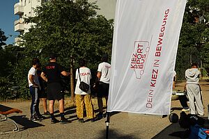 Mehrere junge Menschen, Rückenansicht, stehen draußen vor einem Baum und sprechen miteinander. Sie tragen T-Shirts mit der Aufschrift „team“. Angeschnitten im Bild sind Sportgeräte. Im Vordergrund ein wehendes Banner mit Logo und Aufschrift: „Kiezsportbus. Dein Kiez in Bewegung“, im Hintergrund ein Wohnhaus.