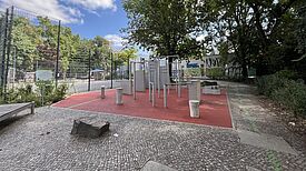 Parkour-Geräte stehen auf einer roten Fläche neben einem eingezäunten Ballspielplatz und Bäumen.