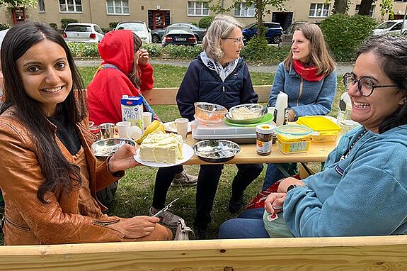 Menschen sitzen beim Dorfcafé im Germaniagarten an einer langen Bank und essen gemeinsam.