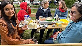 Menschen sitzen beim Dorfcafé im Germaniagarten an einer langen Bank und essen gemeinsam.