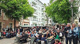 Viele Menschen sitzen und stehen an Tischen zwischen Marktständen auf einer gesperrten, von Bäumen gesäumten Straße.