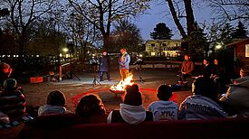 Menschen sitzen im Kreis um eine Feuerschale während Erzählende in der Kreismitte Geschichten vortragen.