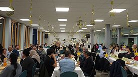 Nachbarinnen und Nachbarn sitzen an langen Tischen in einer Schulmensa und feiern gemeinsam das Fastenbrechen Iftar.