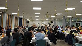 Nachbarinnen und Nachbarn sitzen an langen Tischen in einer Schulmensa und feiern gemeinsam das Fastenbrechen Iftar.