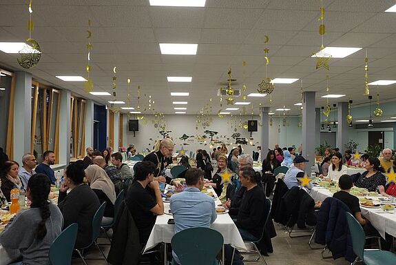 Nachbarinnen und Nachbarn sitzen an langen Tischen in einer Schulmensa und feiern gemeinsam das Fastenbrechen Iftar.