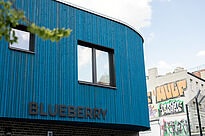 Die Nahaufnahme zeigt die blaue Holzfassade des Blueberry-Gebäudes mit einem Fenster und dem Schriftzug „BLUEBERRY“. Rechts im Hintergrund stehen benachbarte Gebäude, deren Wände mit Graffiti bemalt sind.