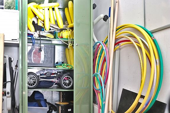 In einem offenen Metallschrank des Spieleverleiiehs liegen gelbe Wurfstäben, bunte Hula-Hoop-Reifen, Holzstangen und ein Kinderwagen.