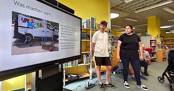Zwei Personen aus dem Team stehen in der Stadtteilbibliothek neben einer Präsentationsfolie mit dem Titel „Was machen wir?“ und erläutern ihr Angebot.