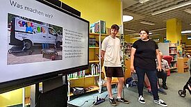 Zwei Personen aus dem Team stehen in der Stadtteilbibliothek neben einer Präsentationsfolie mit dem Titel „Was machen wir?“ und erläutern ihr Angebot.
