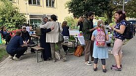 Menschen sitzen und stehen an Tischen im Freien, Kinder spielen, während Pizza am mobilen Kiezofen gebacken wird.
