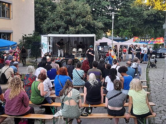 Menschen sitzen und stehen auf einem Platz vor einer kleinen Bühne mit Musikanlage und Schlagzeug. Im Hintergrund befinden sich Bäume und ein Banner, auf dem Fête de la Musique steht.