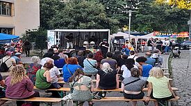 Menschen sitzen und stehen auf einem Platz vor einer kleinen Bühne mit Musikanlage und Schlagzeug. Im Hintergrund befinden sich Bäume und ein Banner, auf dem Fête de la Musique steht.