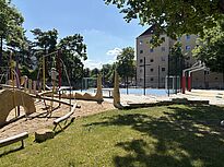 Im Vordergrund verläuft eine Rasenfläche im Schatten der Bäume. Dahinter steht ein Spielbereich mit Sandboden, Balancierstangen und Kletterelementen, während rechts daneben ein eingezäunter Bolzplatz mit Basketballkörben liegt.