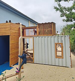 Eine Konstruktion im Außenraum verbindet einen begehbaren Holzkasten mit einem begehbaren Container. Auf dem grauen Container mit Fenster und Durchgang ist ein kleiner Holztunnel zum Durchkriechen bis in den Holzkasten gebaut. Kinder spielen im Container und auf Kletterbalken davor. Heller, weicher Untergrund, im Hintergrund grüne Sträucher, links eine hellblaue Wand.