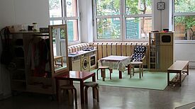Heller Kita-Raum mit Spielküche, kleinen Tischen und Stühlen.
