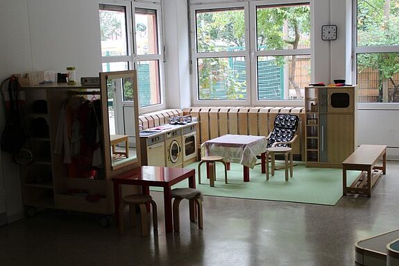Heller Kita-Raum mit Spielküche, kleinen Tischen und Stühlen.