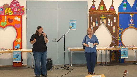 Zwei Frauen stehen bei der Superschüler*innen-Ehrung 2025 im QM-Gebiet Rollbergsiedlung vor einer bunt dekorierten Wand und sprechen ins Mikrofon.