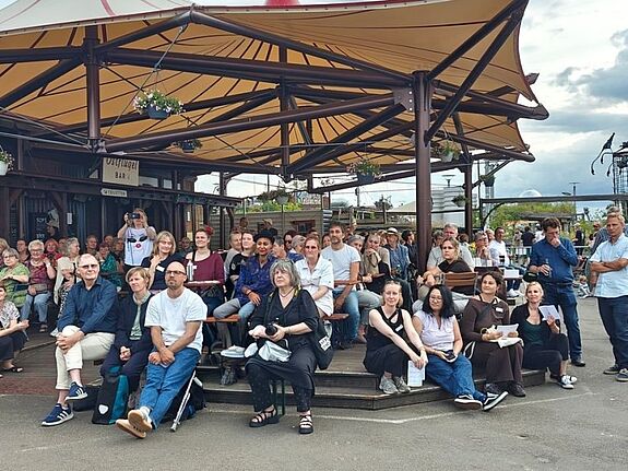 Viele Engagierte sitzen und stehen auf dem Parkdeck des Klunkerkranichs unter einem großen Sonnensegel und nehmen am Sommerfest teil. 