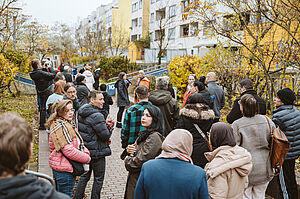 Auf einem Weg zwischen einer bepflanzten Rabatte und Wohnungsneubauten stehen viele Menschen. Sie sprechen miteinander und schauen sich um, einige fotografieren. 
