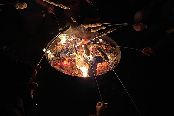 Menschen backen Stockbrot über einer Feuerschale, die in der Dunkelheit leuchtet.