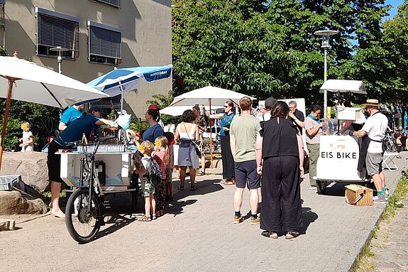 Menschen stehen bei Sonnenschein an Ständen mit Sonnenschirmen, einer Wasserbar und einem Eisfahrrad auf dem Sasarsteig, während Kinder sich Eis holen.