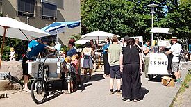 Menschen stehen bei Sonnenschein an Ständen mit Sonnenschirmen, einer Wasserbar und einem Eisfahrrad auf dem Sasarsteig, während Kinder sich Eis holen.