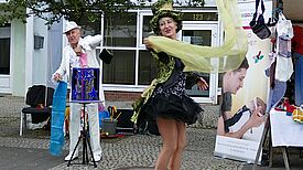 Eine Frau mit Zauberhut und in einem kurzen, grün-schwarzen Zauberkostüm tanzt mit einem grünen Tuch in der Hand auf der Straße. Im Hintergrund steht ein Zauberer und zieht weitere Tücher aus einer Schachtel.