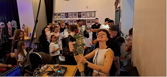 Frau mit Blumenstrauß im Vordergrund und zahlreiche Menschen im Hintergrund im Backstage-Bereich der Modenschau im Kulturzentrum Staaken.