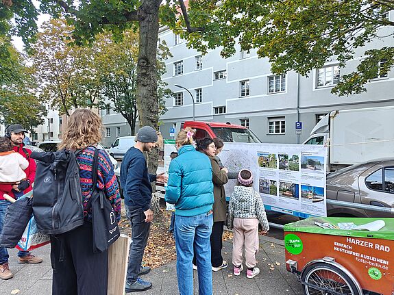 Erwachsene und ein Kind betrachten auf dem Gehweg eine Infotafel mit Karte und Begrünungsbeispielen.