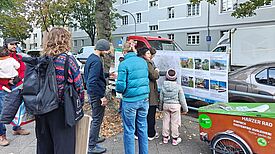 Erwachsene und ein Kind betrachten auf dem Gehweg eine Infotafel mit Karte und Begrünungsbeispielen.