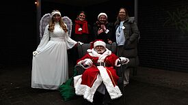 Nikolaus sitzt im Sessel, Engel und QM-Teammitglieder stehen daneben auf dem Wintermarkt in der Thermometersiedlung.